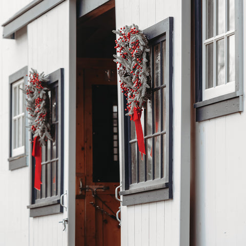 Decorative wreaths with red ribbons hanging on a white barn with black-framed windows.