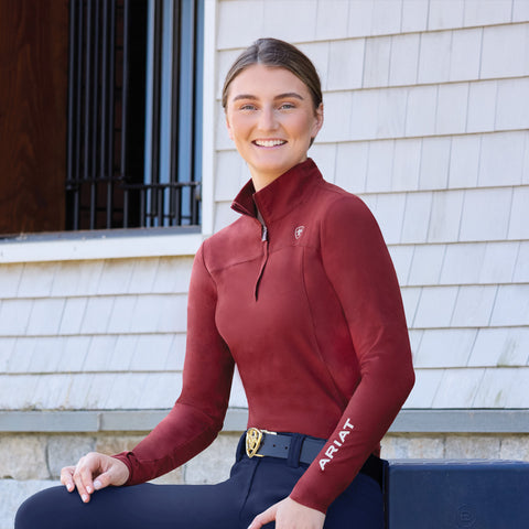 Woman wearing a red equestrian shirt with 'Ariat' branding, sitting against a white wall.